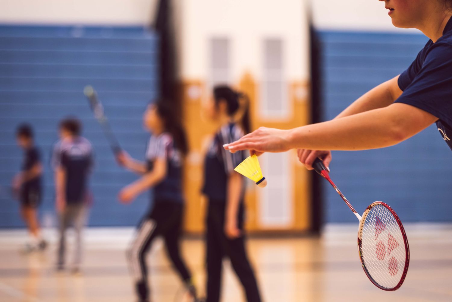 A player preparing a badminton serve while others practise in a sports hall